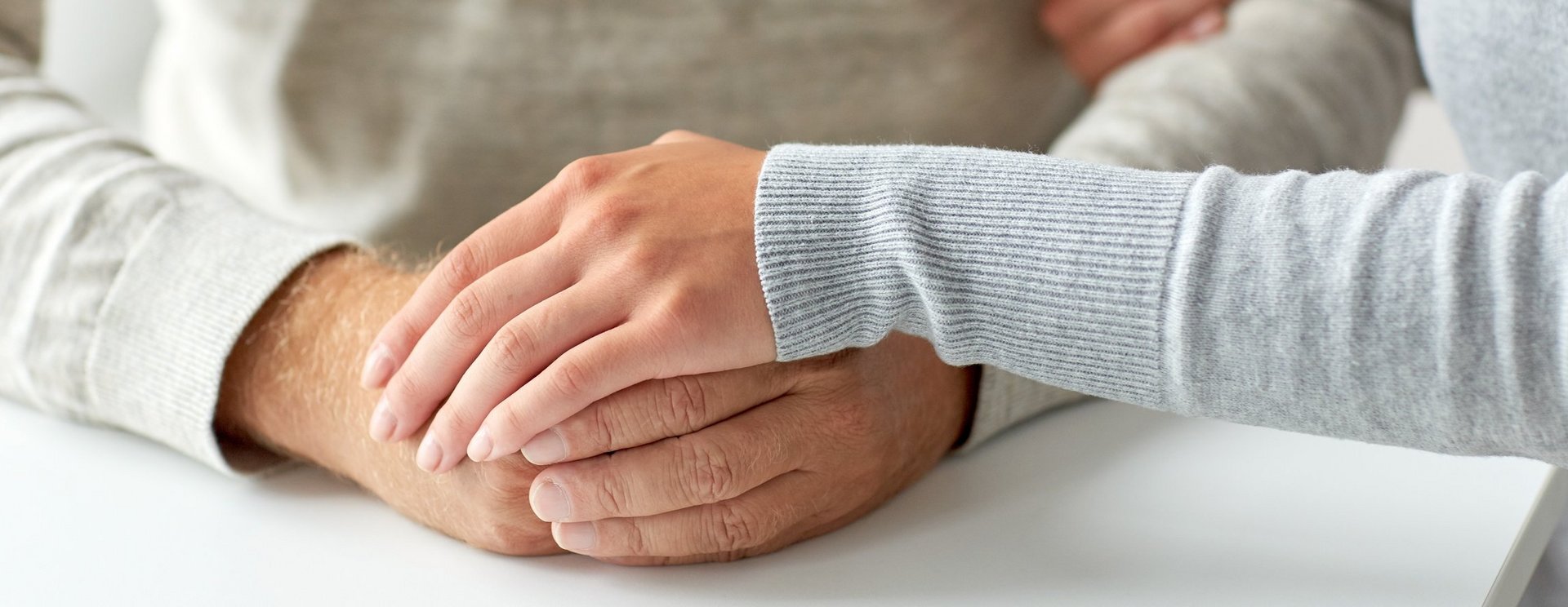 Ein Mann legt seine gefaltete Hände auf einen Tisch und eine Frau legt ihr Hand behütet auf die gefalteten Hände.