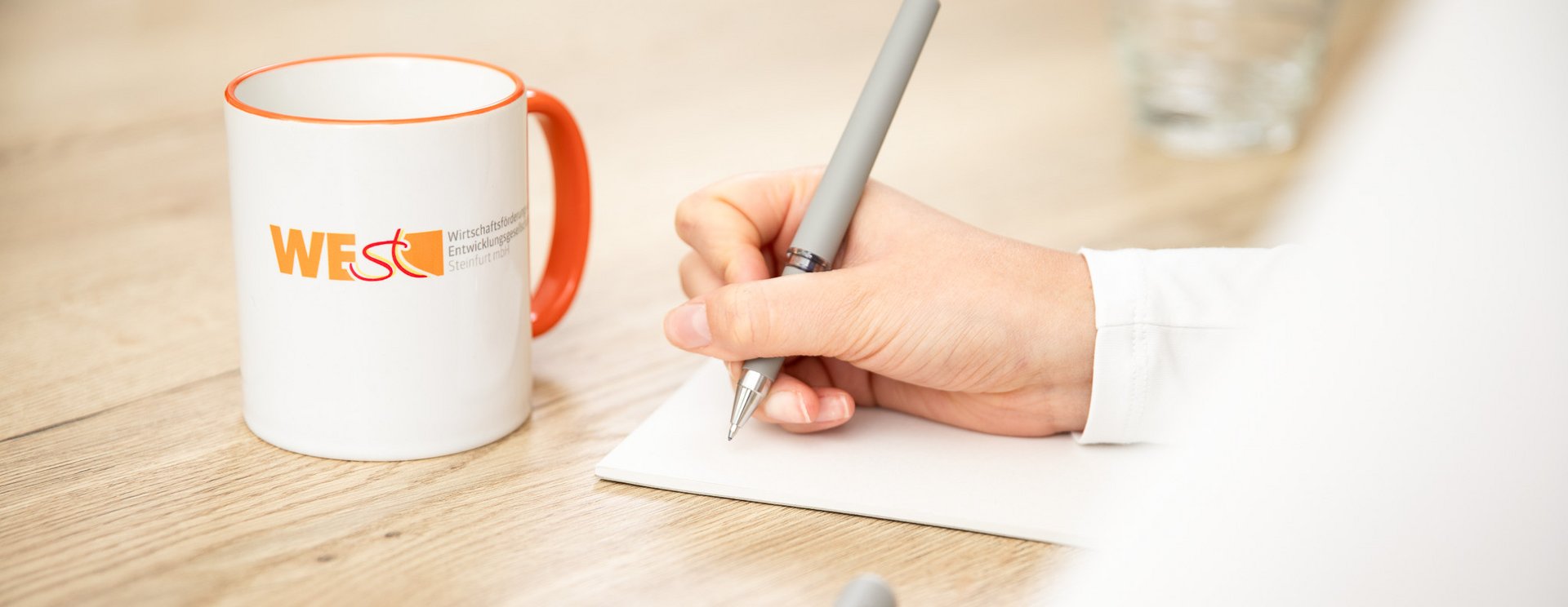 Auf einem Tisch steht eine Tasse mit dem Schriftzug "WESt". Daneben liegt ein Block auf dem jemand etwas notiert.