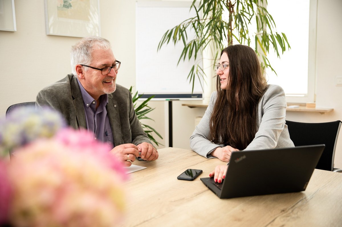 Zwei Personen sitzen an einem Tisch und unterhalten sich, eine Person hat einen Laptop vor sich.