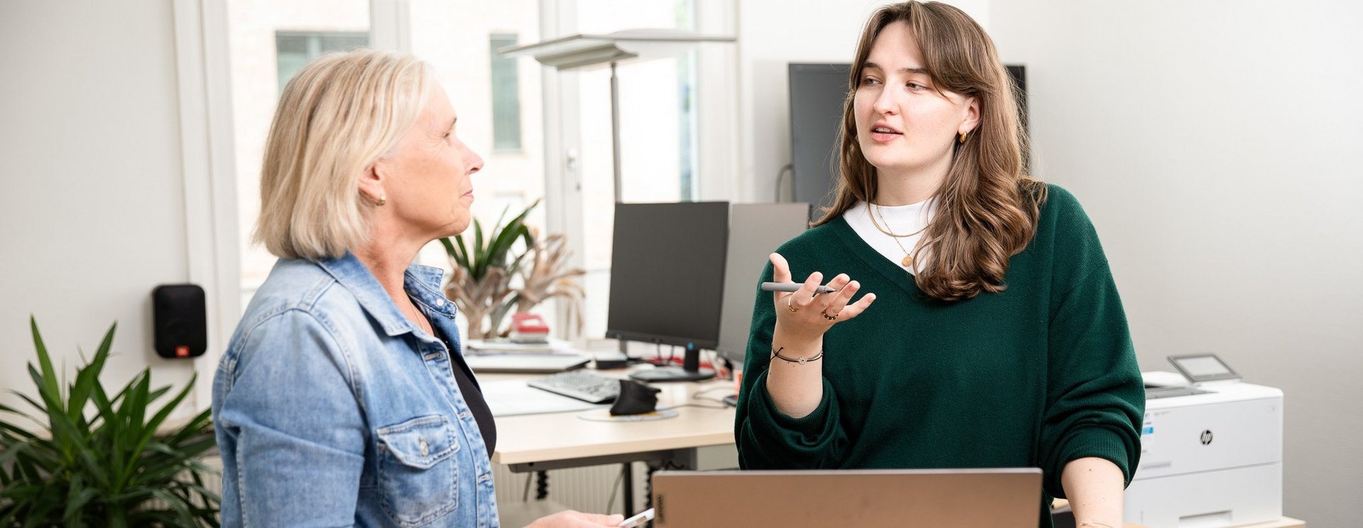 Zwei Frauen unterhalten sich in einem Büro. Vor Ihnen steht ein geöffneter Laptop.