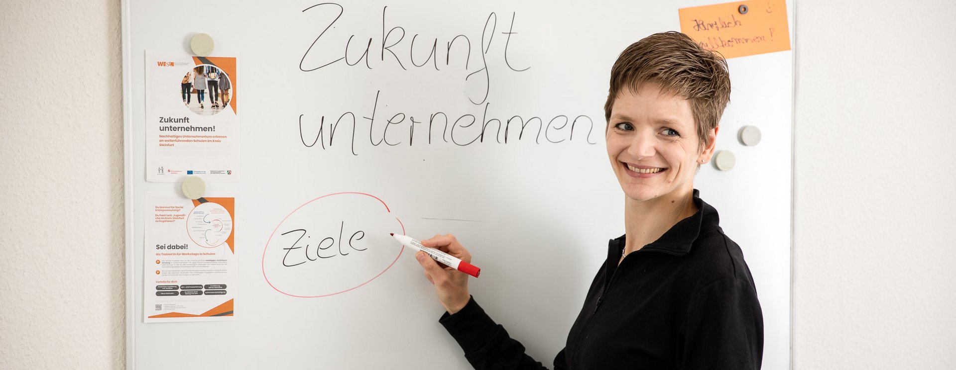 Frau steht an einem WhiteBoard und schreibt den Text "Zukunft unternehmen!" sowie das Wort "Ziele". Sie schaut lächelnd in die Kamera.