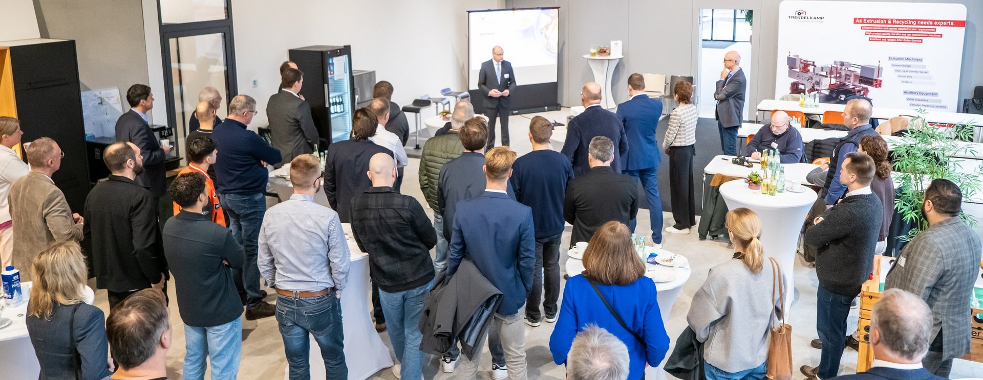 Ein großes Publikum steht in einem modernen Foyer und hört einem Vortrag zu.