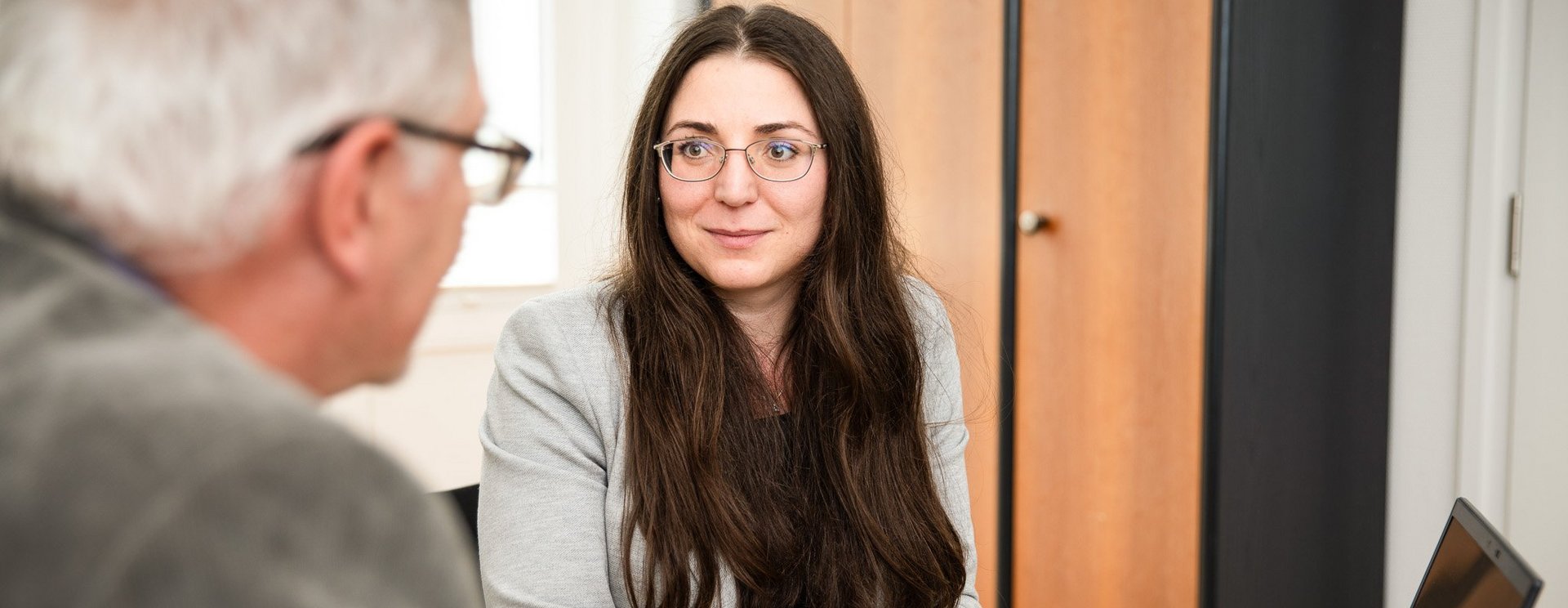 Eine Person sitzt vor einem Laptop und unterhält sich mit einer weiteren Person, die im Anschnitt zu sehen ist.