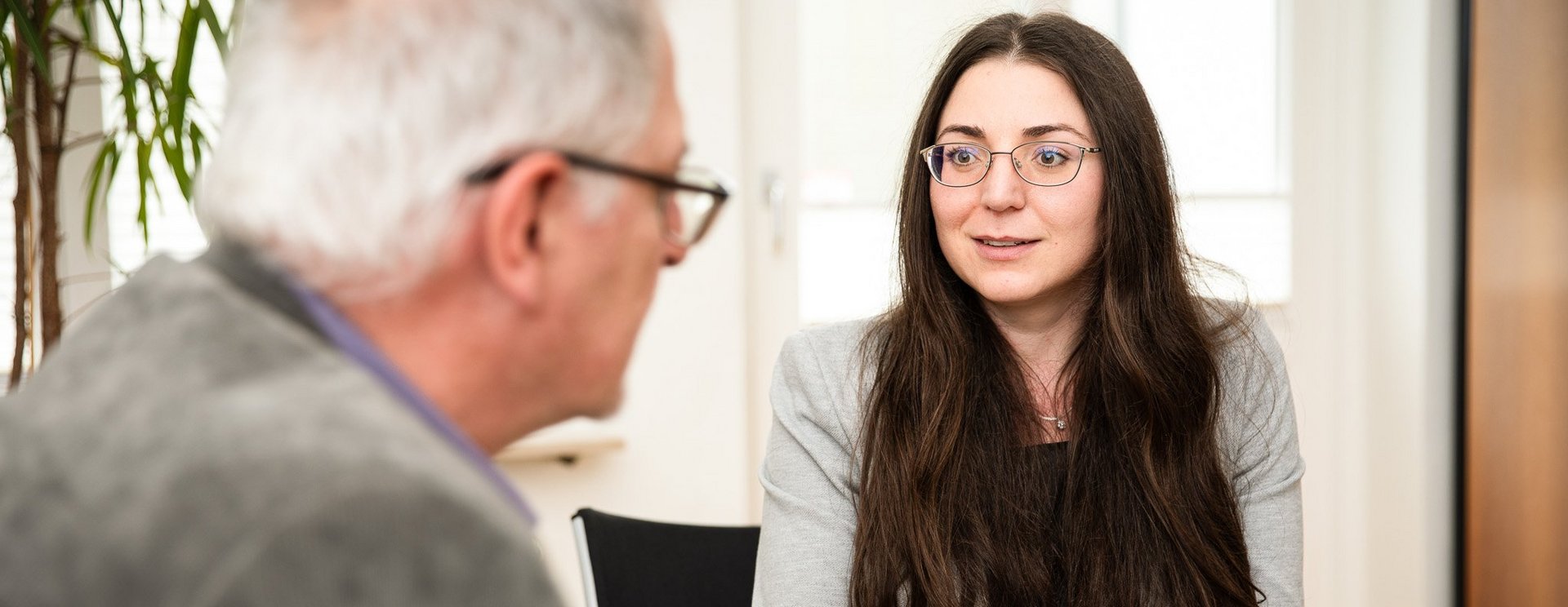 Eine Person sitzt vor einem Laptop und unterhält sich mit einer weiteren Person, die im Anschnitt zu sehen ist.