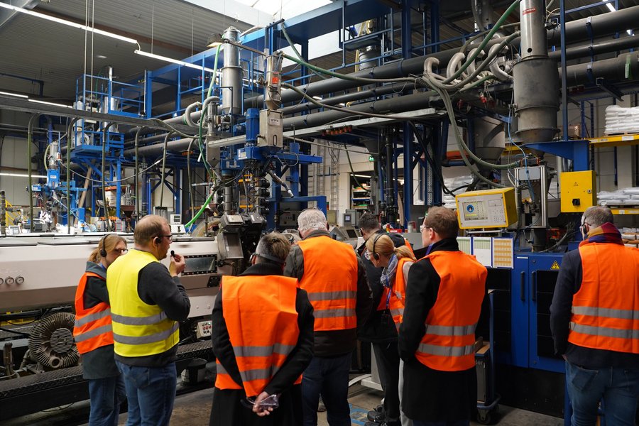 Eine Gruppe von Menschen in orangenen Warnwesten stehen in einer industriellen Produktionshalle.