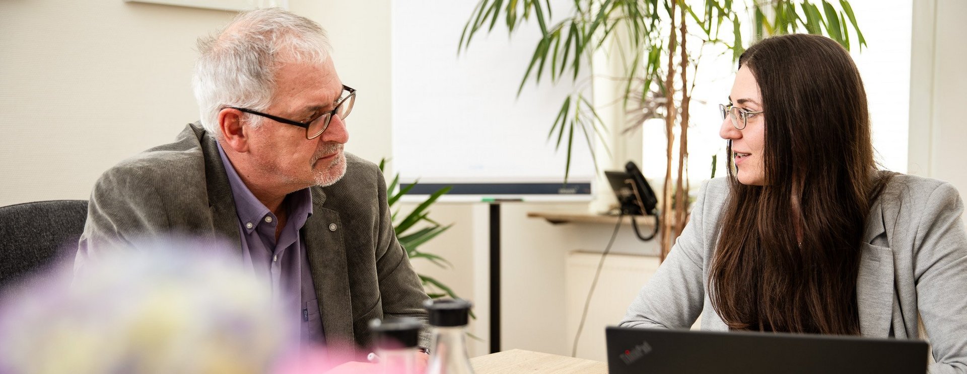 Zwei Personen sitzen an einem Tisch und unterhalten sich, eine Person hat einen Laptop vor sich.