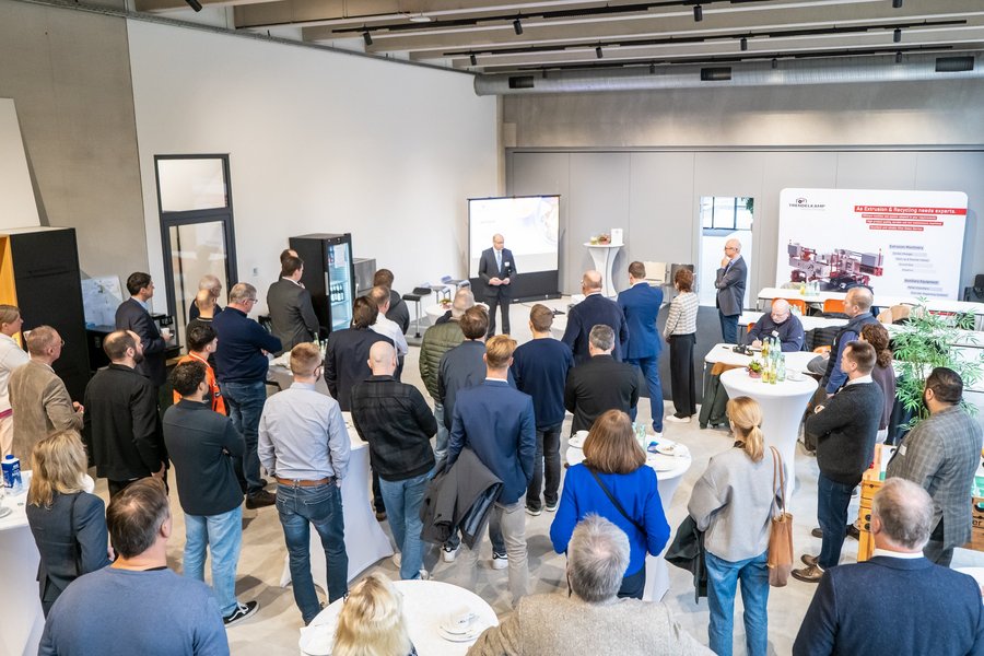 Ein großes Publikum steht in einem modernen Foyer und hört einem Vortrag zu.