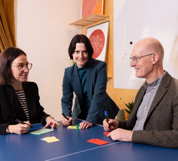 3 Personen sitzen an einem blauen Tisch und halten bunte Kärtchen in der Hand.