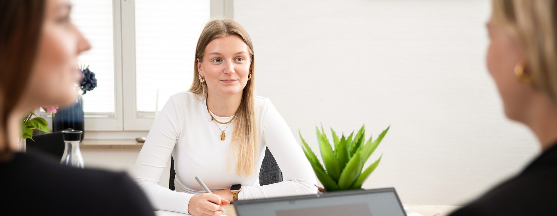 Drei Personen sitzen an einem Besprechungstisch und unterhalten sich, eine Person sitzt vor einem Laptop.