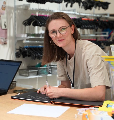 Eine Frau steht in einem Laden, schreibt etwas in ein Buch und lächelt in die Kamera.