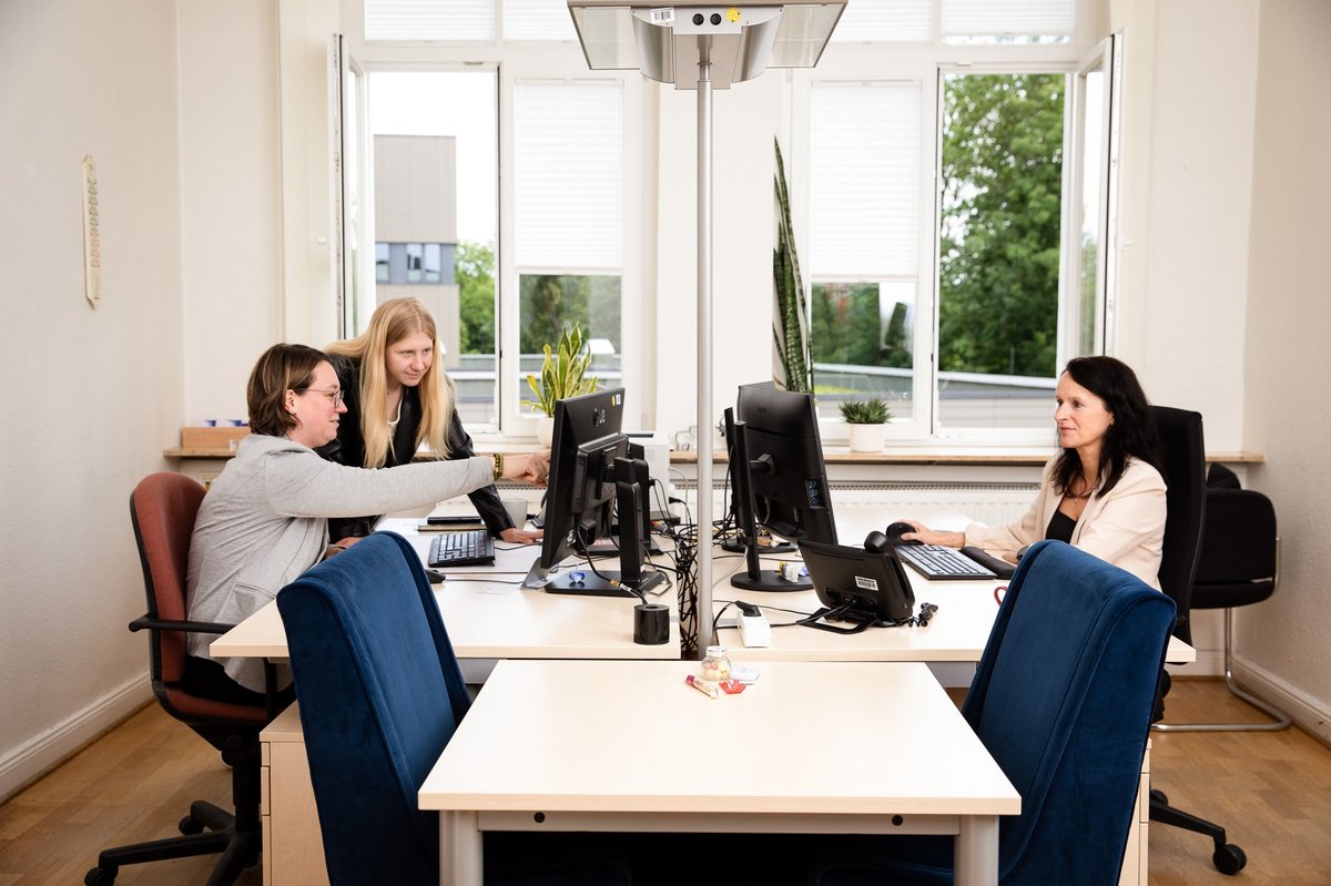 Drei Personen sitzen sich in einem Büro gegenüber, 2 von ihnen schauen gemeinsam auf einen Bildschirm.